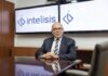 Intelisis: La evolución del ERP; de la gestión de información a la toma de decisiones Fotografía de retrato corporativo en plano medio de Dionisio Castillo Guillén, Director General de Intelisis Software, vistiendo traje oscuro con corbata a rayas y lentes, sentado a la mesa de una sala de juntas frente a dos pantallas con el logotipo de Intelisis.