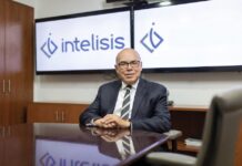 Intelisis: La evolución del ERP; de la gestión de información a la toma de decisiones Fotografía de retrato corporativo en plano medio de Dionisio Castillo Guillén, Director General de Intelisis Software, vistiendo traje oscuro con corbata a rayas y lentes, sentado a la mesa de una sala de juntas frente a dos pantallas con el logotipo de Intelisis.