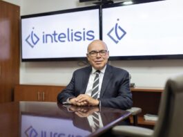 Intelisis: La evolución del ERP; de la gestión de información a la toma de decisiones Fotografía de retrato corporativo en plano medio de Dionisio Castillo Guillén, Director General de Intelisis Software, vistiendo traje oscuro con corbata a rayas y lentes, sentado a la mesa de una sala de juntas frente a dos pantallas con el logotipo de Intelisis.