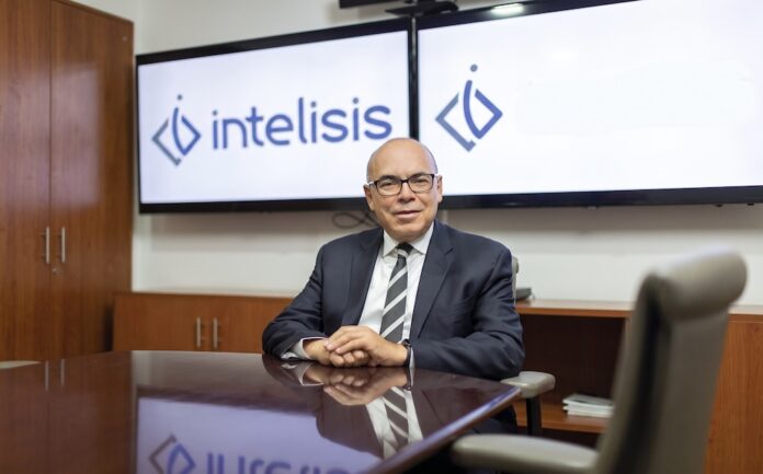 Dionisio Castillo Guillén reflexiona sobre los 40 años de evolución de los sistemas ERP en México y América Latina Fotografía de retrato corporativo en plano medio de Dionisio Castillo Guillén, Director General de Intelisis Software, vistiendo traje oscuro con corbata a rayas y lentes, sentado a la mesa de una sala de juntas frente a dos pantallas con el logotipo de Intelisis.
