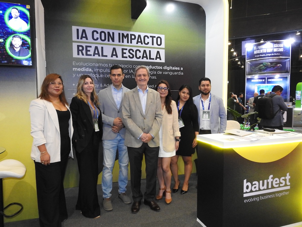 Fotografía de grupo en plano entero de siete ejecutivos y consultores del equipo de Baufest posando sonrientes en su estand corporativo junto al mostrador principal y frente al letrero "IA CON IMPACTO REAL A ESCALA".