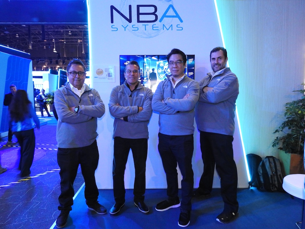 Fotografía de grupo en plano entero de cuatro ingenieros y ejecutivos de NBA Systems vistiendo suéteres grises corporativos idénticos, posando sonrientes con los brazos cruzados frente al muro publicitario iluminado de su estand de exhibición.
