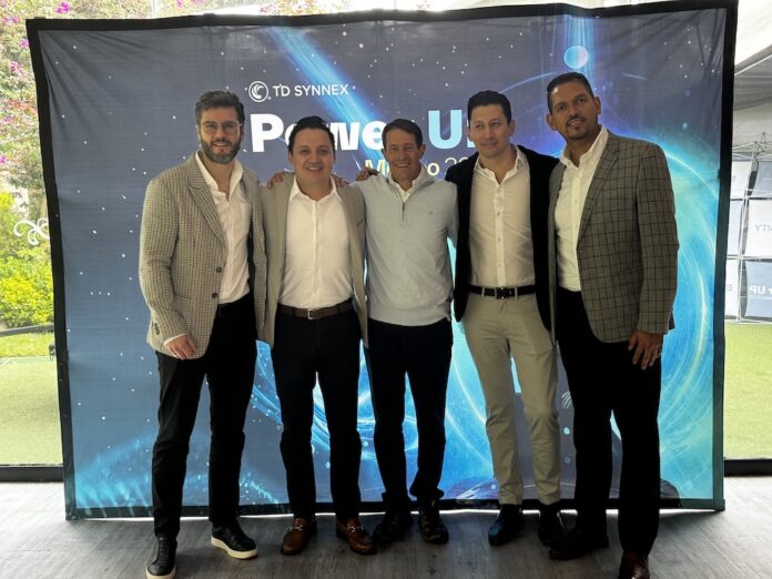 Fotografía de grupo de los cinco directivos principales de TD SYNNEX México —Jaime Venegas, Gerardo Calderón, Raffaelo Piccolo, Fabián Gómez y José Luis Chávez— posando abrazados y sonrientes frente a una mampara publicitaria azul de Power Up México 2026.