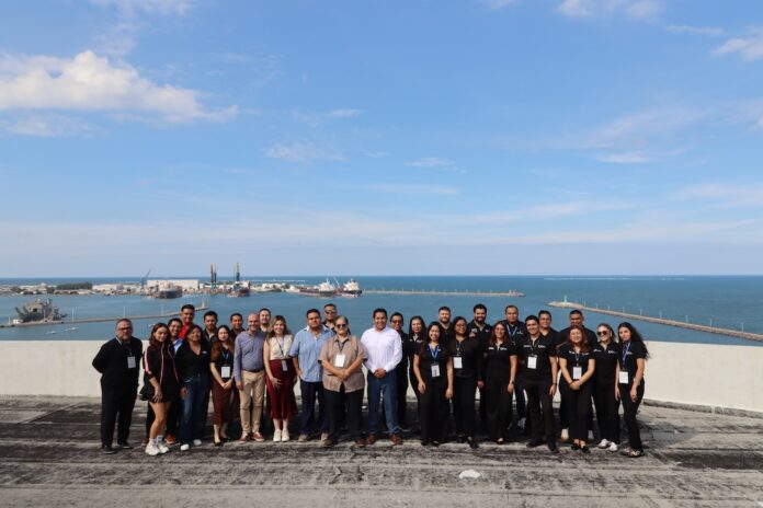 Fotografía grupal en plano entero del equipo organizador de Grupo CVA posando sonrientes en una terraza ejecutiva con el puerto y el mar de Veracruz al fondo.