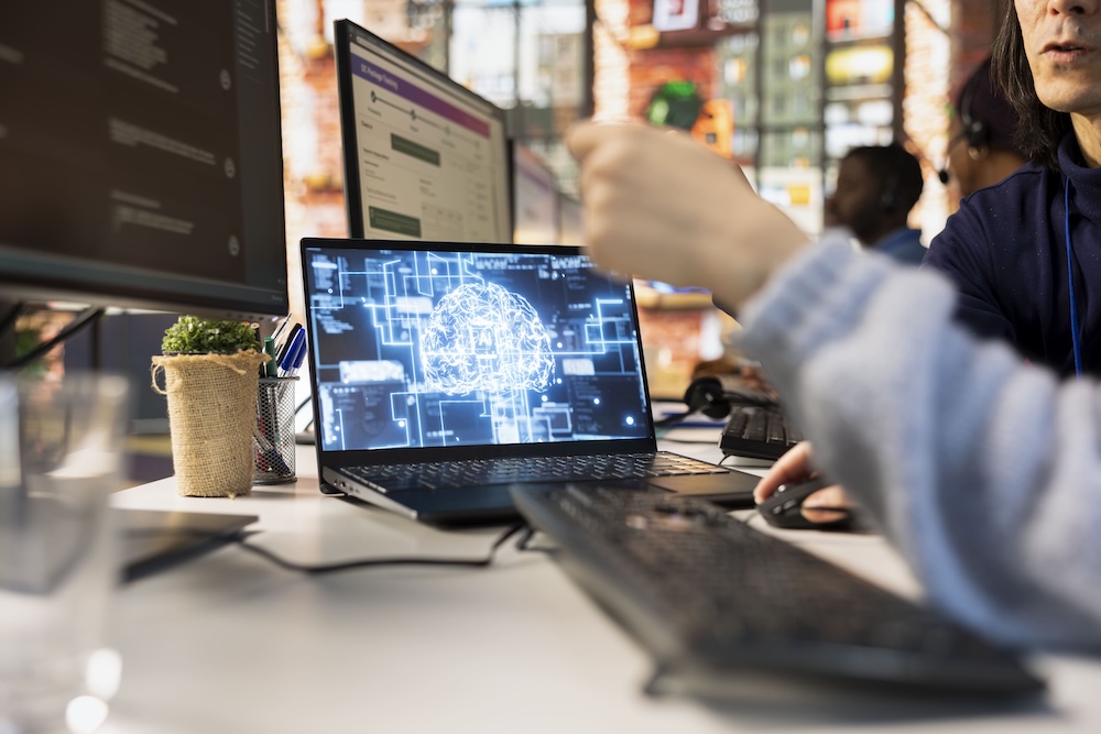 Fotografía en plano detalle dentro de una oficina colaborativa donde un empleado señala hacia la pantalla de una computadora portátil abierta que muestra un cerebro digital con circuitos y nodos de datos interconectados en azul eléctrico.