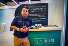 IKUSI redefine la madurez digital con arquitecturas de próxima generación Fotografía en plano medio de Ernesto Gutiérrez, Especialista en redes, infraestructura TI y observabilidad en IKUSI, vistiendo camisa polo azul marino corporativa y posando apoyado en un mostrador promocional de IKUSI con la leyenda conmemorativa de 55 años.