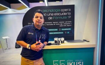 IKUSI redefine la madurez digital con arquitecturas de próxima generación Fotografía en plano medio de Ernesto Gutiérrez, Especialista en redes, infraestructura TI y observabilidad en IKUSI, vistiendo camisa polo azul marino corporativa y posando apoyado en un mostrador promocional de IKUSI con la leyenda conmemorativa de 55 años.