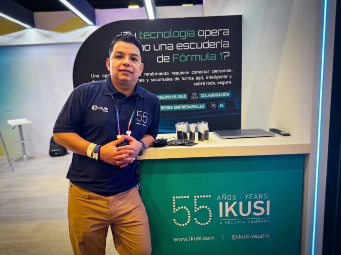 Fotografía en plano medio de Ernesto Gutiérrez, Especialista en redes, infraestructura TI y observabilidad en IKUSI, vistiendo camisa polo azul marino corporativa y posando apoyado en un mostrador promocional de IKUSI con la leyenda conmemorativa de 55 años.