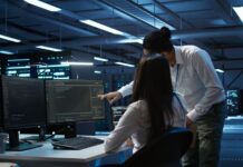 Zebra Technologies: Ciberseguridad estratégica para proteger la cadena de suministro en México Fotografía en plano medio de una ingeniera de sistemas sentada frente a múltiples monitores analizando líneas de código y un especialista de TI inclinado a su lado señalando la pantalla en una sala de servidor o centro de datos moderno iluminado con luces azules.