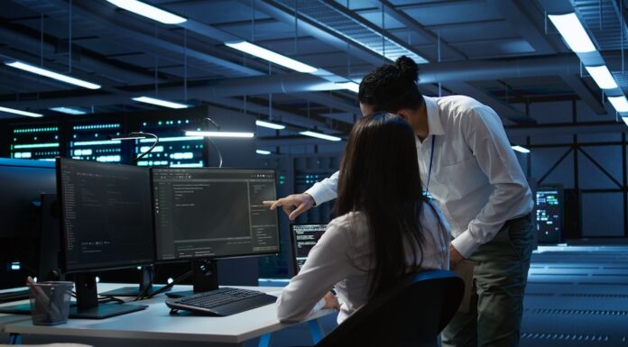 Zebra Technologies: Ciberseguridad estratégica para proteger la cadena de suministro en México Fotografía en plano medio de una ingeniera de sistemas sentada frente a múltiples monitores analizando líneas de código y un especialista de TI inclinado a su lado señalando la pantalla en una sala de servidor o centro de datos moderno iluminado con luces azules.