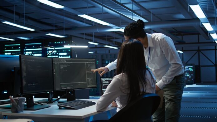 Especialistas de TI monitorean la seguridad de la red corporativa y la infraestructura en un centro de datos Fotografía en plano medio de una ingeniera de sistemas sentada frente a múltiples monitores analizando líneas de código y un especialista de TI inclinado a su lado señalando la pantalla en una sala de servidor o centro de datos moderno iluminado con luces azules.