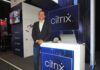 Citrix redefine el futuro laboral con una adopción segura de inteligencia artificial Fotografía en plano medio de Fabio Charry, Partner Territory Manager para Centroamérica, México y el Caribe en Citrix, vistiendo un saco oscuro sobre camisa blanca y jeans, posando sonriente junto a un mostrador iluminado con el logotipo azul de Citrix.