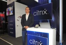 Citrix redefine el futuro laboral con una adopción segura de inteligencia artificial Fotografía en plano medio de Fabio Charry, Partner Territory Manager para Centroamérica, México y el Caribe en Citrix, vistiendo un saco oscuro sobre camisa blanca y jeans, posando sonriente junto a un mostrador iluminado con el logotipo azul de Citrix.
