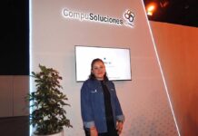 El futuro tecnológico se consolida con la exitosa alianza entre CompuSoluciones y Dell Technologies Fotografía en plano medio de Fabiola Munguía de pie en una camisa de mezclilla azul con el logotipo de CompuSoluciones, posando sonriente junto a una planta en maceta frente al muro de exhibición de la marca.