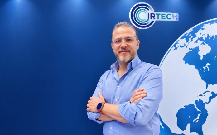 Fotografía de retrato en plano medio de Fernando Rondero, Director de ciberseguridad y tecnología en Grupo Cirtec, vistiendo una camisa azul claro, posando sonriente con los brazos cruzados frente a un muro azul corporativo con el logotipo de Cirtec y la ilustración de un mapa mundi.
