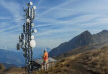 GlobalLogic lanza un framework con IA agéntica para transformar las pruebas en redes de banda ancha y 5G Fotografía en plano general de un técnico de telecomunicaciones con casco blanco y chaleco reflejante naranja de espaldas, sosteniendo un dispositivo digital frente a una torre de antenas y repetidoras en la cima de una montaña bajo el cielo azul.