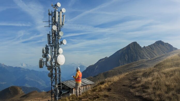 Fotografía en plano general de un técnico de telecomunicaciones con casco blanco y chaleco reflejante naranja de espaldas, sosteniendo un dispositivo digital frente a una torre de antenas y repetidoras en la cima de una montaña bajo el cielo azul.