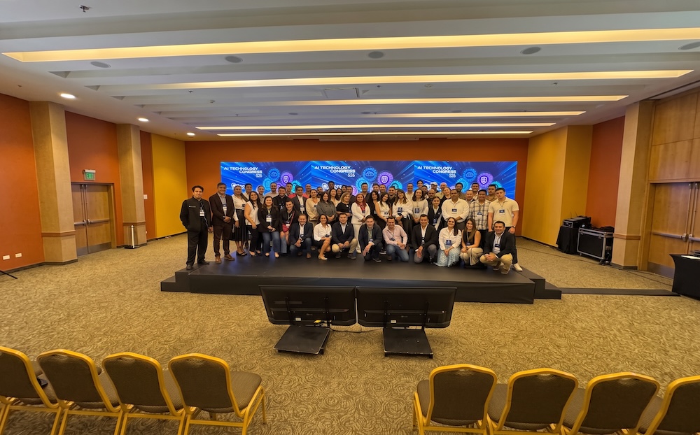 Fotografía de grupo en plano entero de todos los participantes, organizadores y conferencistas posando sonrientes sobre el escenario del salón de eventos frente a las pantallas corporativas del evento.
