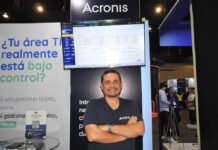 Acronis y el salto hacia la ciberseguridad impulsada por inteligencia artificial en Latinoamérica Fotografía de cuerpo medio de Guillermo Márquez, Solution Engineer Manager para Latinoamérica en Acronis, sonriendo con los brazos cruzados con una camiseta negra corporativa en un estand de exhibición frente a un monitor que muestra la interfaz de la plataforma y un cartel con la frase "¿Tu área TI realmente está bajo control?".