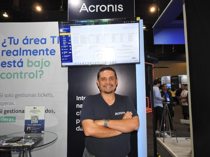 Guillermo Márquez destaca las soluciones de ciberseguridad e IA de Acronis para América Latina Fotografía de cuerpo medio de Guillermo Márquez, Solution Engineer Manager para Latinoamérica en Acronis, sonriendo con los brazos cruzados con una camiseta negra corporativa en un estand de exhibición frente a un monitor que muestra la interfaz de la plataforma y un cartel con la frase
