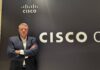 Cisco redefine las oportunidades y los retos para los socios de negocio en América Latina Fotografía de retrato en plano medio de Guy Diedrich, Senior VP y Global Innovation Officer de Cisco, vistiendo saco oscuro sobre camisa blanca, posando sonriente y con los brazos cruzados frente al muro con el logotipo corporativo de CISCO.