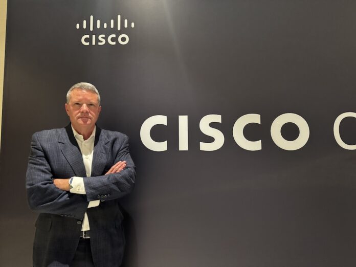 Fotografía de retrato en plano medio de Guy Diedrich, Senior VP y Global Innovation Officer de Cisco, vistiendo saco oscuro sobre camisa blanca, posando sonriente y con los brazos cruzados frente al muro con el logotipo corporativo de CISCO.