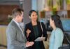 HPE impulsa a sus partners en México con mayor certidumbre comercial y mejores incentivos Fotografía en plano medio de tres ejecutivos de negocios conversando sonrientes en el vestíbulo de una oficina moderna, donde una mujer al centro vistiendo saco negro gesticula mientras sostiene una libreta junto a un colega con saco gris y otra ejecutiva con saco tejido verde menta.