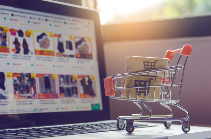 Hostinger revela que la preparación anticipada impulsa hasta un 36.8% los pedidos online durante El Buen Fin Carrito de compras miniatura con detalles rojos que transporta dos paquetes de cartón sobre el teclado de una computadora portátil abierta, mostrando en la pantalla desenfocada un catálogo de comercio electrónico con productos y descuentos.