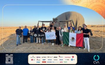 Grupo CVA fortalece su ecosistema tecnológico desde Marruecos e impulsa una nueva etapa de innovación Fotografía grupal en plano entero de ejecutivos y fabricantes posando sonrientes frente a un globo aerostático posado en el suelo del desierto, sosteniendo la bandera de México y la bandera corporativa de CVA.