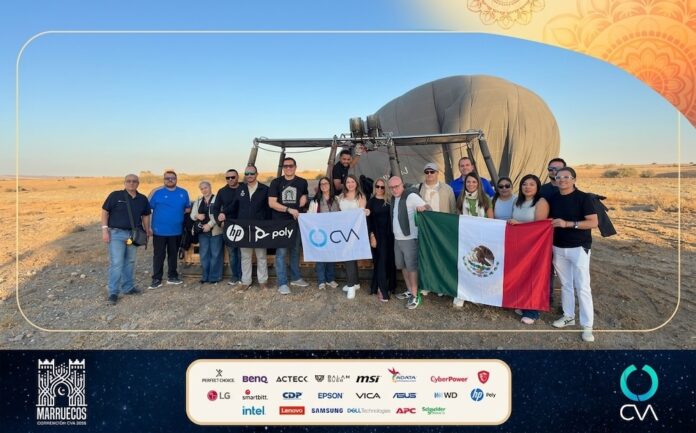 Canales de distribución y patrocinadores exhiben la bandera mexicana junto a la bandera de CVA tras una actividad grupal en Marruecos. Fotografía grupal en plano entero de ejecutivos y fabricantes posando sonrientes frente a un globo aerostático posado en el suelo del desierto, sosteniendo la bandera de México y la bandera corporativa de CVA.