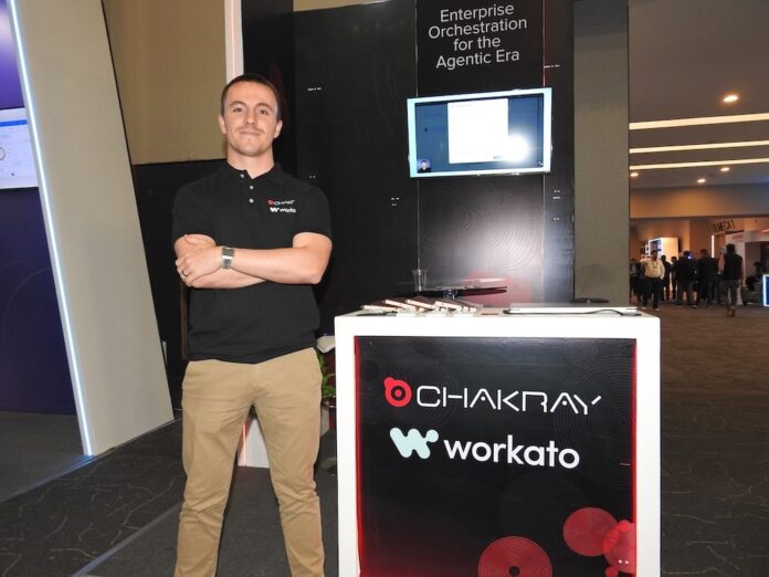 Fotografía en plano entero de Jon García, Líder de la práctica de Workato en Chakray, posando con los brazos cruzados vistiendo una playera polo negra con los bordados corporativos de Chakray y Workato, junto al estand de exhibición.