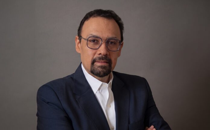 Fotografía de retrato en plano medio de Jorge López, Vicepresidente de Tanium Latinoamérica, vistiendo un saco azul marino sobre una camisa blanca de vestir, con anteojos y barba de candado, posando con los brazos cruzados frente a un fondo neutro gris.