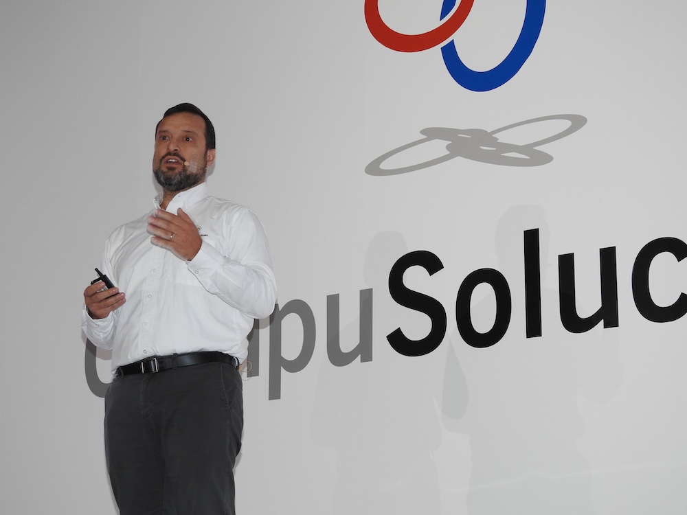 Fotografía en plano medio de Jos Medina Mora, Director Internacional en CompuSoluciones, vistiendo camisa blanca, gesticulando durante su presentación frente al logotipo gigante de la empresa.