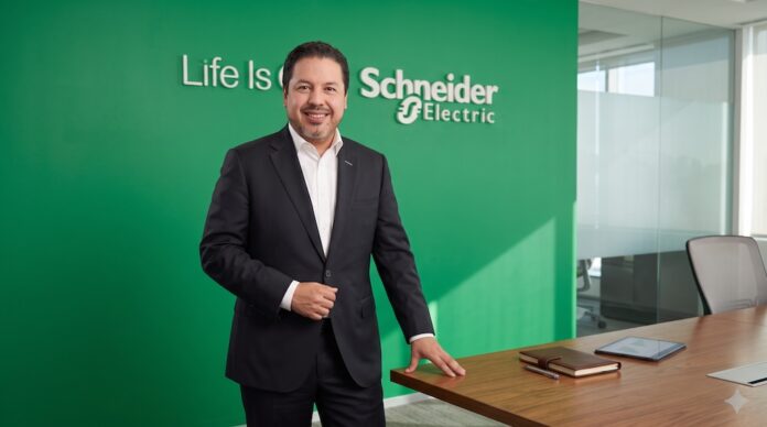 José Alberto Llavot destaca el diseño modular y la flexibilidad en centros de datos para Inteligencia Artificial Fotografía en plano medio de José Alberto Llavot, Gerente de Preventa y Desarrollador de Negocios en Schneider Electric para México y Centroamérica, vistiendo saco oscuro sobre camisa blanca, posando sonriente y apoyado en una mesa de madera de sala de juntas frente a un muro verde con el logotipo corporativo iluminado.