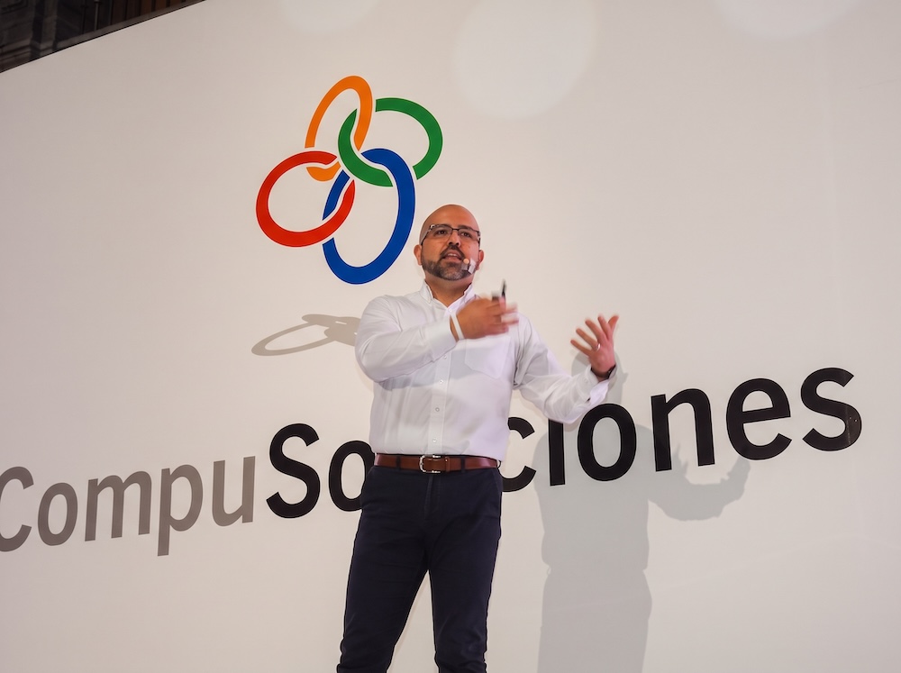 Fotografía en plano medio de José Luis Cortés, Director Comercial de CompuSoluciones, gesticulando sonriente sobre el escenario frente al logotipo mural iluminado del mayorista.