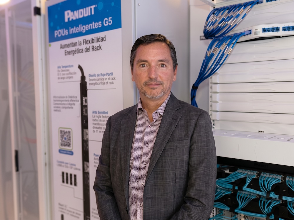 Fotografía en plano medio de Juan Pablo Borray, Sr. Business Development Manager Latam para Panduit, vistiendo un saco a cuadros gris sobre camisa de vestir, posando junto a un rack de telecomunicaciones con cableado estructurado azul organizadamente conectado.