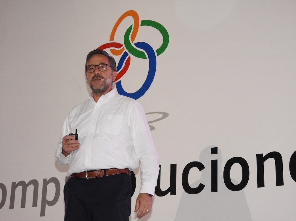 Fotografía en plano medio de Juan Pablo Medina Mora, Presidente Ejecutivo de CompuSoluciones, vistiendo camisa blanca de vestir con anteojos y micrófono de solapa, exponiendo de pie frente al logotipo mural corporativo.