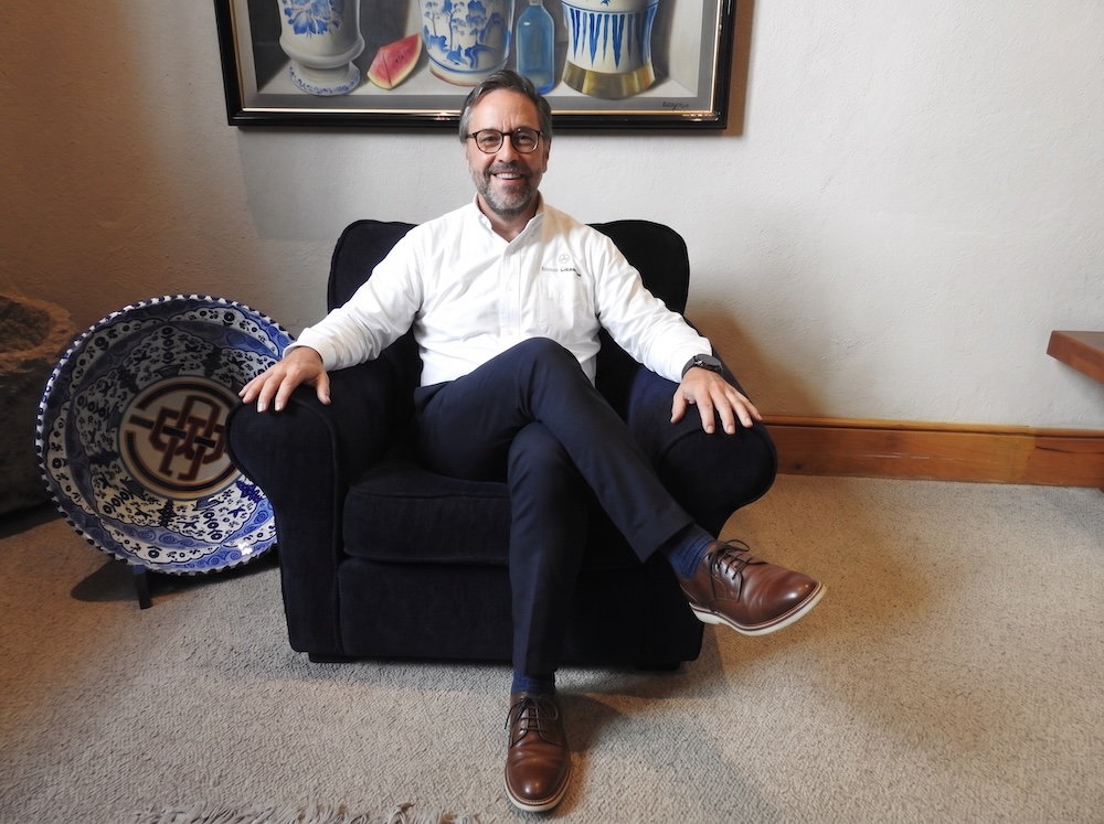 Fotografía en plano entero de Juan Pablo Medina Mora, Presidente Ejecutivo de CompuSoluciones, vistiendo camisa blanca corporativa y anteojos, sentado sonriente con las piernas cruzadas en un sillón oscuro.