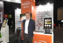 El futuro de la integración digital avanza con Belltech Fotografía en plano medio de Juan Pedro Esquerré, Country Manager de Belltech para Chile y México, vistiendo un saco azul marino sobre una camisa blanca de vestir, posando frente a un estand corporativo decorado con el logotipo naranja de Belltech y flanqueado por quioscos digitales de autoatención con pantalla táctil.
