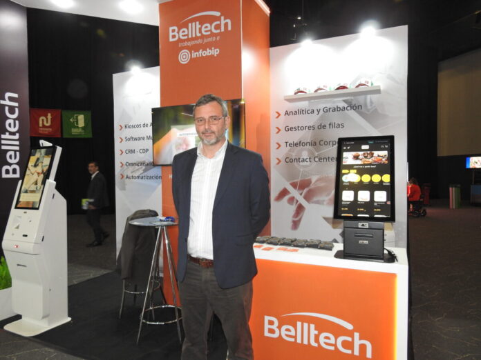 Fotografía en plano medio de Juan Pedro Esquerré, Country Manager de Belltech para Chile y México, vistiendo un saco azul marino sobre una camisa blanca de vestir, posando frente a un estand corporativo decorado con el logotipo naranja de Belltech y flanqueado por quioscos digitales de autoatención con pantalla táctil.