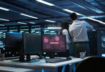 KIO IT Services advierte sobre la nueva era de ciberataques con IA y los cinco riesgos rumbo a 2027 Fotografía en plano general de un centro de operaciones de seguridad (SOC) o sala de servidores en penumbra, donde dos analistas de espaldas revisan los racks mientras en el monitor de un escritorio en primer plano se muestra una alerta roja con el mensaje "SYSTEM HACKED".