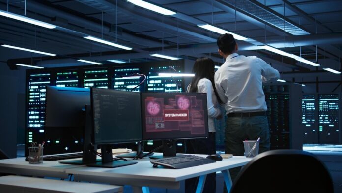 Fotografía en plano general de un centro de operaciones de seguridad (SOC) o sala de servidores en penumbra, donde dos analistas de espaldas revisan los racks mientras en el monitor de un escritorio en primer plano se muestra una alerta roja con el mensaje 