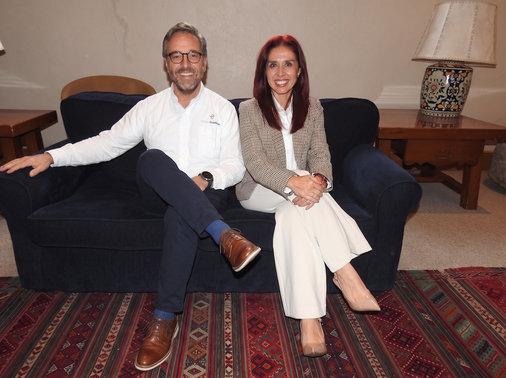 Fotografía en plano medio de Juan Pablo Medina Mora y Ana María Arreola posando juntos y sonrientes sentados en un sillón azul en las instalaciones corporativas de CompuSoluciones.