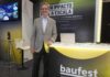 El impacto real de la inteligencia artificial en los negocios según la visión de Baufest Fotografía en plano medio de Luis Battilana, Country Manager de Baufest, sonriendo en un saco claro sobre una camisa de vestir azul, apoyado en el mostrador iluminado del estand de Baufest bajo el lema "IA CON IMPACTO REAL A ESCALA".