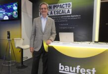 El impacto real de la inteligencia artificial en los negocios según la visión de Baufest Fotografía en plano medio de Luis Battilana, Country Manager de Baufest, sonriendo en un saco claro sobre una camisa de vestir azul, apoyado en el mostrador iluminado del estand de Baufest bajo el lema "IA CON IMPACTO REAL A ESCALA".