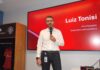 Orquestando el futuro digital: El poder de la IA Agéntica de Snapdragon Fotografía en plano medio de Luiz Tonisi, VP y Presidente de Qualcomm Latinoamérica, vistiendo camisa blanca de botones y pantalón negro, hablando al micrófono frente a una pantalla roja corporativa que muestra su nombre y cargo.