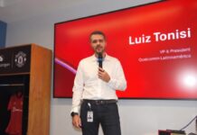 Orquestando el futuro digital: El poder de la IA Agéntica de Snapdragon Fotografía en plano medio de Luiz Tonisi, VP y Presidente de Qualcomm Latinoamérica, vistiendo camisa blanca de botones y pantalón negro, hablando al micrófono frente a una pantalla roja corporativa que muestra su nombre y cargo.