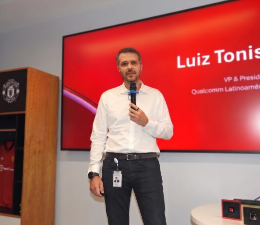 Orquestando el futuro digital: El poder de la IA Agéntica de Snapdragon Fotografía en plano medio de Luiz Tonisi, VP y Presidente de Qualcomm Latinoamérica, vistiendo camisa blanca de botones y pantalón negro, hablando al micrófono frente a una pantalla roja corporativa que muestra su nombre y cargo.