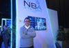El aliado tecnológico corporativo y el impacto transformador de NBA Systems Fotografía en plano medio de Marco González, Director de NBA Systems, vistiendo un suéter gris corporativo bordado sobre una camisa blanca, posando con los brazos cruzados frente al estand con pantalla interactiva y logotipo iluminado de la empresa.