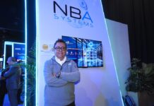 El aliado tecnológico corporativo y el impacto transformador de NBA Systems Fotografía en plano medio de Marco González, Director de NBA Systems, vistiendo un suéter gris corporativo bordado sobre una camisa blanca, posando con los brazos cruzados frente al estand con pantalla interactiva y logotipo iluminado de la empresa.